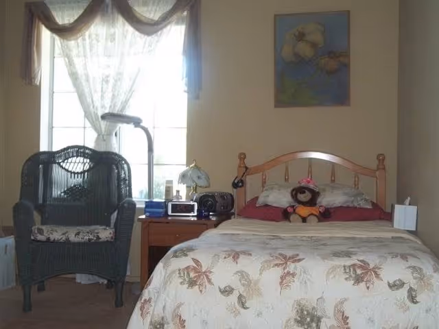 A cozy bedroom with a wooden bed frame, floral patterned bedspread, and a teddy bear wearing a pink hat and orange outfit sitting on the bed. Next to the bed is a wooden nightstand with a lamp, clock radio, and other small items. A black wicker chair with a floral cushion is positioned near a window with sheer white curtains and a decorative valance. A floral painting hangs on the wall above the bed.