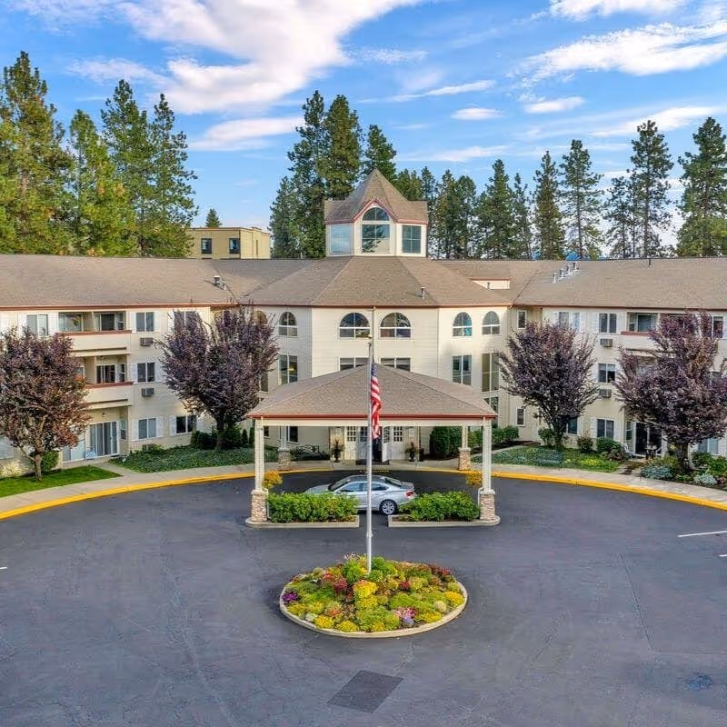 Front entrance of a multi-story senior living facility with a covered drive-through, flagpole and landscaped circular driveway.