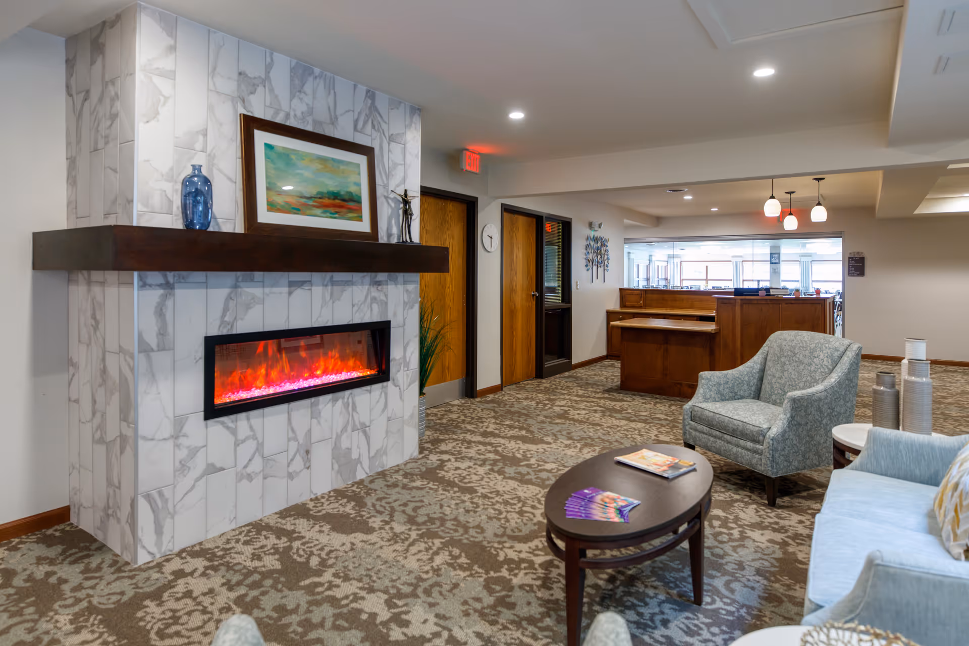 A cozy living room area in a senior living facility featuring a modern electric fireplace with a marble-patterned surround and a wooden mantel. On the mantel are decorative items including a blue vase and a framed painting. The room has patterned carpet flooring, a light blue upholstered armchair, a matching sofa, and a dark wooden coffee table with magazines and brochures. In the background, there is a wooden reception desk and doors leading to other rooms, with ceiling lights providing warm illumination.