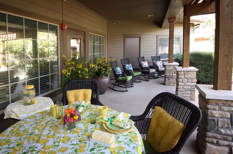Covered outdoor patio area with a table set for four, featuring a lemon-patterned tablecloth, yellow cushions on black wicker chairs, a floral centerpiece, and a beverage dispenser. In the background, there are several rocking chairs with colorful cushions lined up against the wall, large potted plants, and stone pillars supporting the roof.