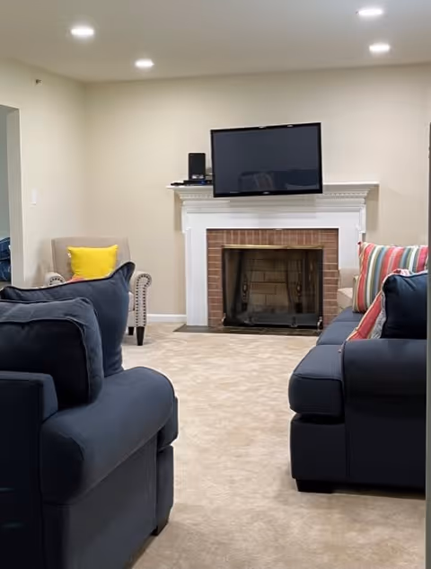 A cozy living room with beige carpet, two dark blue sofas with colorful pillows, a beige armchair with a yellow pillow, a white fireplace with a brick interior, and a flat-screen TV mounted above the fireplace.