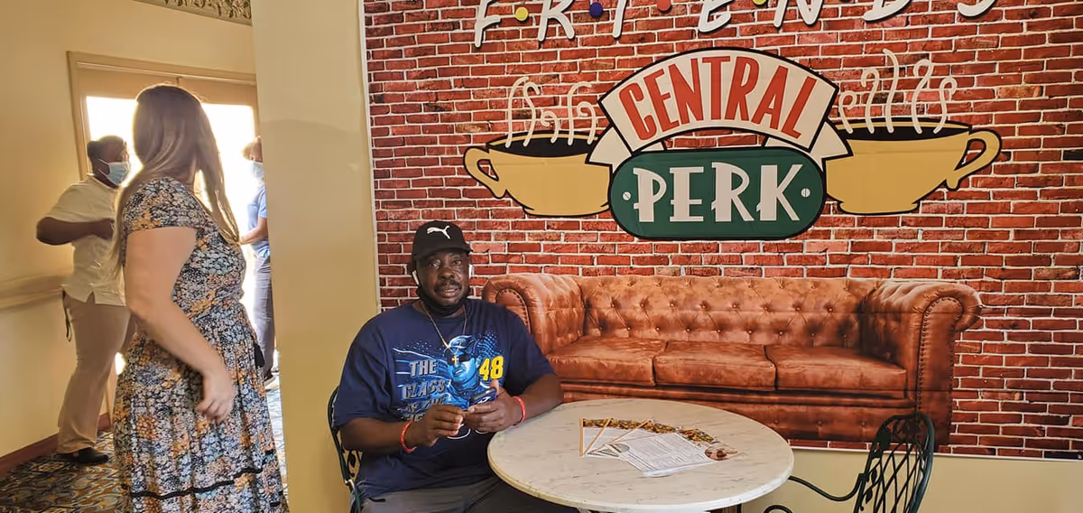 Several people gather in an interior common area decorated with a 'Central Perk' brick-wall mural, a brown tufted couch, and a round table.