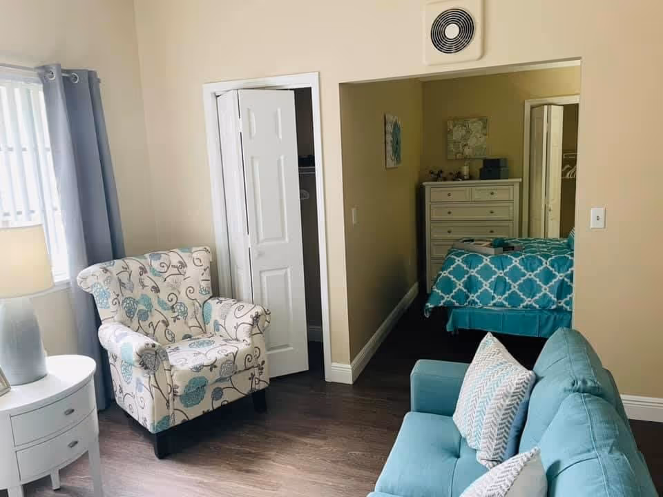Sunlit living area with a patterned armchair and teal sofa, looking into a nearby bedroom with a teal-patterned bedspread.