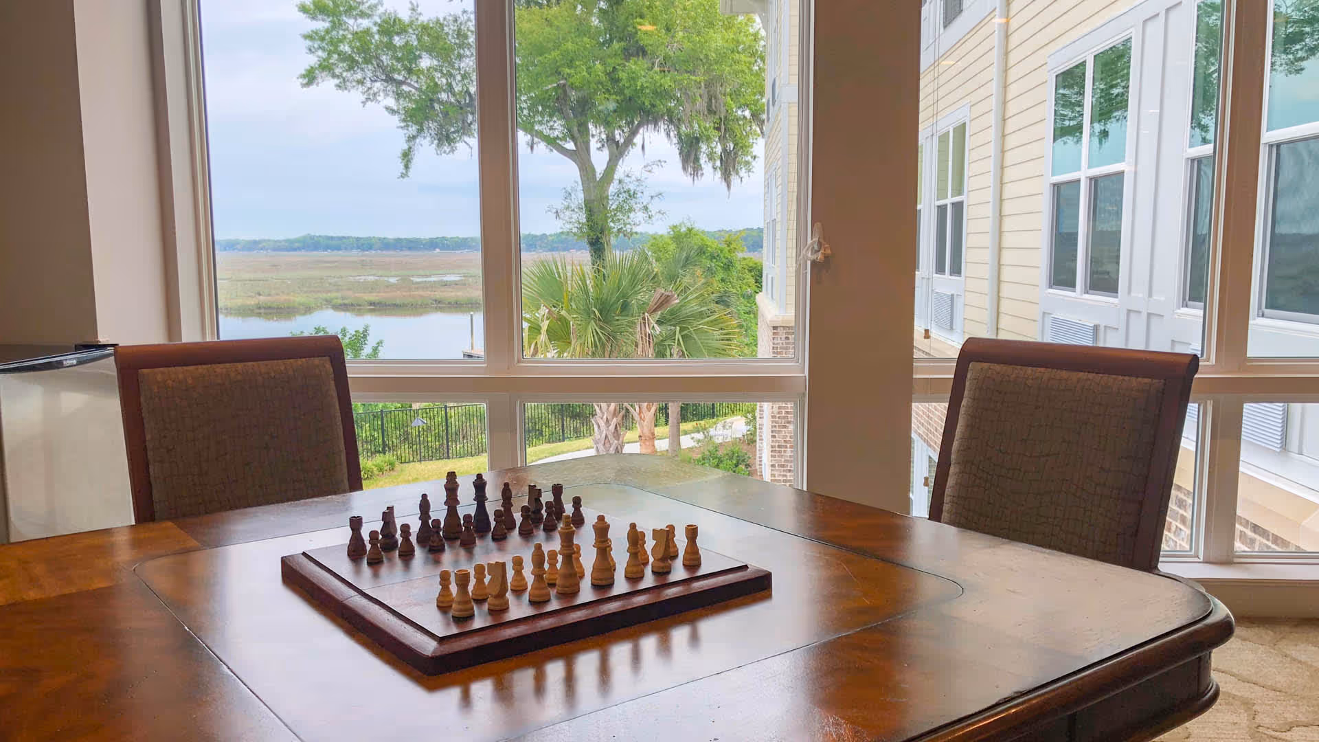 A wooden table with a chessboard and chess pieces set up, surrounded by two upholstered chairs. Large windows behind the table offer a view of trees, a grassy area, and a body of water in the distance.