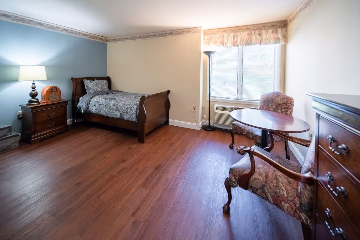 A senior living facility bedroom with a single wooden bed with patterned bedding, a wooden nightstand with a lamp and a vintage radio, two floral upholstered chairs around a small wooden table, a wooden dresser, a floor lamp, and a window with blinds and a valance. The room has wood flooring and light-colored walls with a decorative border near the ceiling.