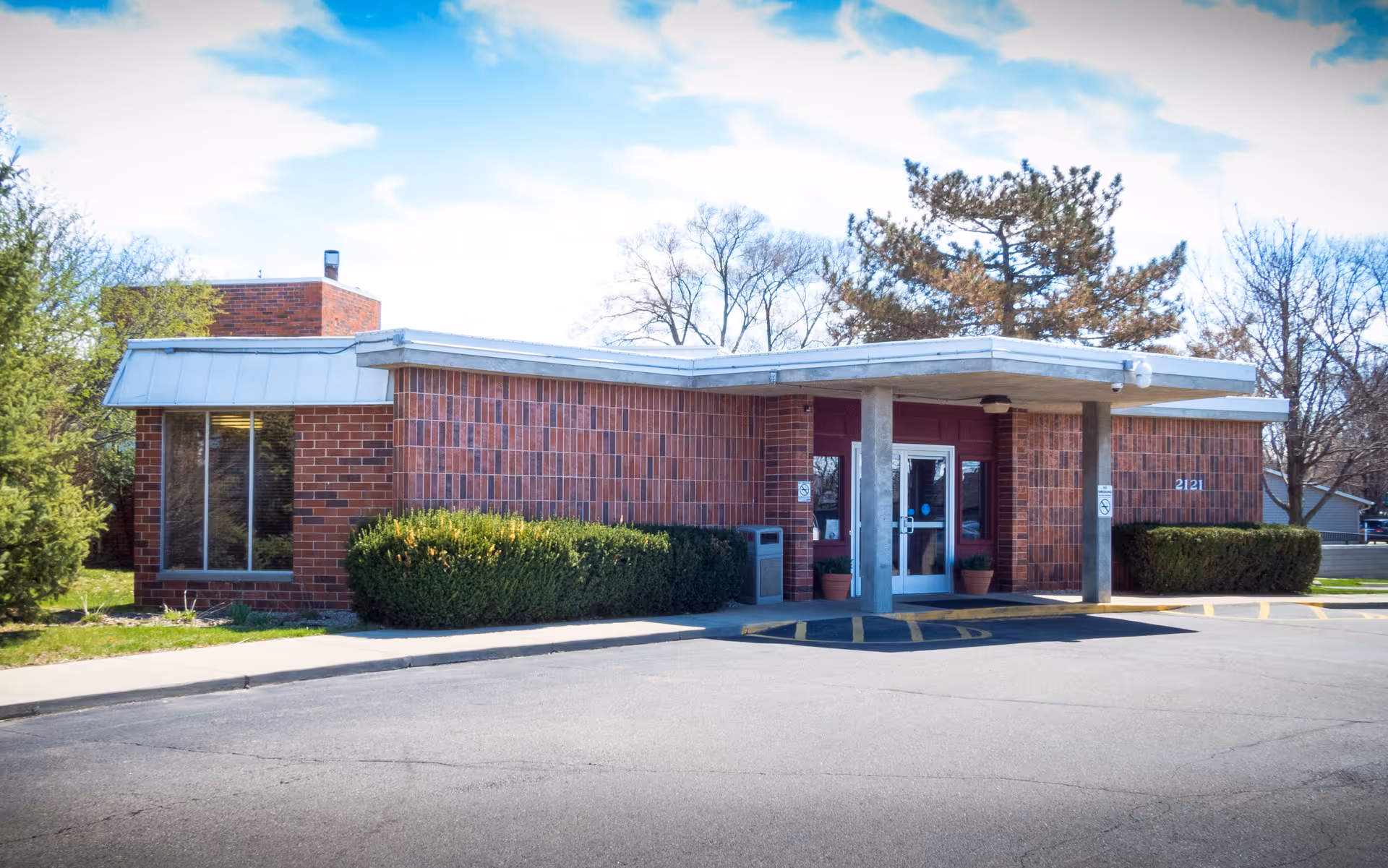 Exterior view of a single-story brick building with a flat roof and a covered entrance supported by two columns. There are bushes along the front of the building and a sidewalk leading to the entrance. The sky is partly cloudy and trees are visible in the background.