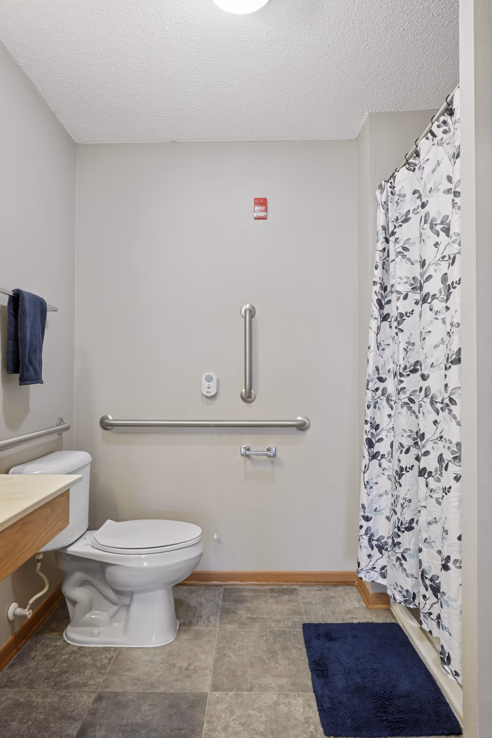 Accessible bathroom with a toilet, horizontal and vertical grab bars, a sink counter, a shower with a floral curtain, and a navy bath mat.