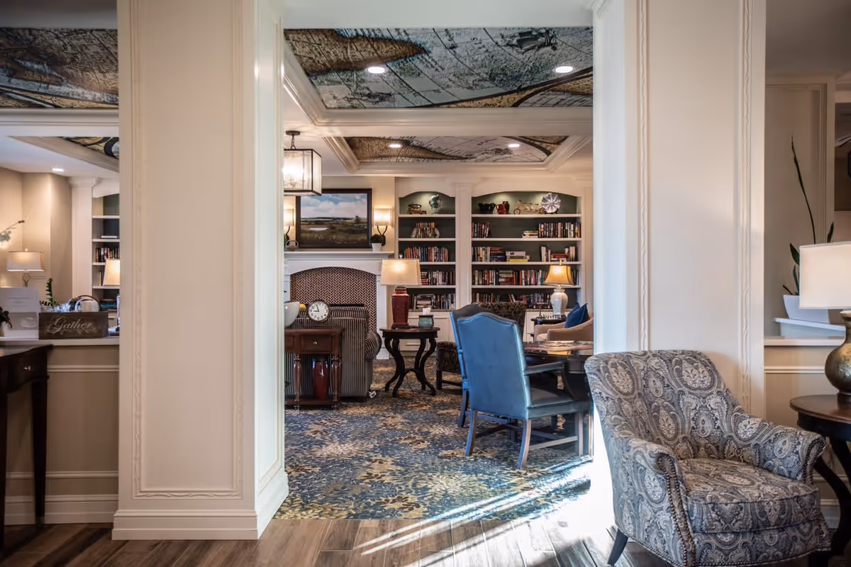 Interior view of a senior living facility common area featuring a cozy seating arrangement with patterned armchairs and blue chairs around a table. The room has built-in bookshelves filled with books and decorative items, a fireplace with a painting above it, and soft lighting from table lamps and wall sconces. The ceiling has artistic designs, and the floor is covered with a patterned carpet transitioning to wood flooring.