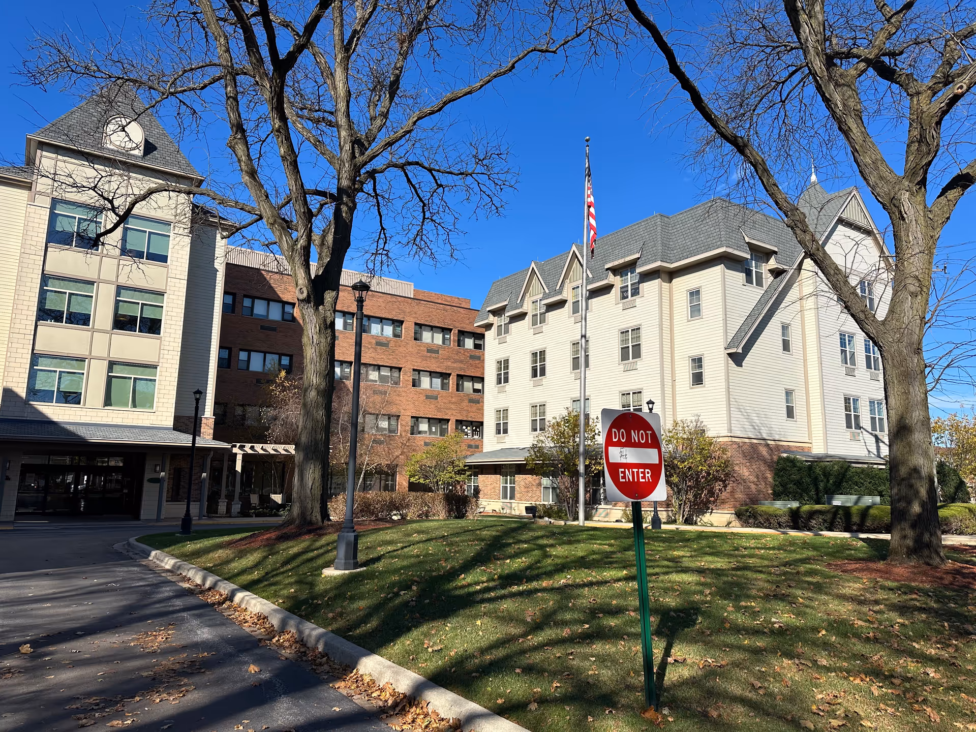 Exterior view of Norwood Crossing facility showing a multi-story building with a mix of brick and light-colored siding. There are large leafless trees in the foreground, a well-maintained lawn, a flagpole with an American flag, and a 'Do Not Enter' sign near the driveway entrance. The sky is clear and blue.