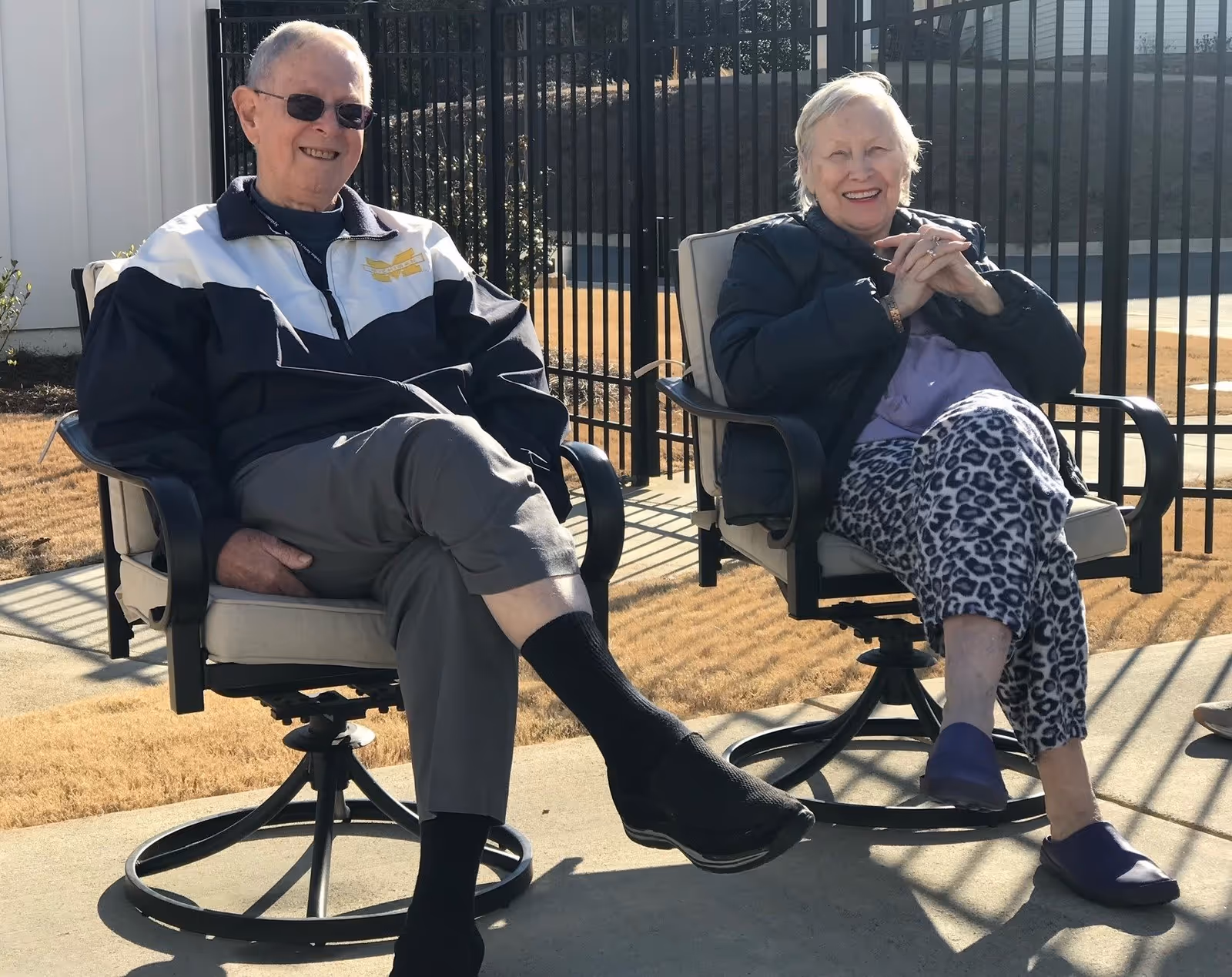An elderly man and woman sitting outdoors on cushioned swivel chairs, smiling and enjoying the sunny weather. They are seated on a concrete patio with a black metal fence and dry grass in the background.