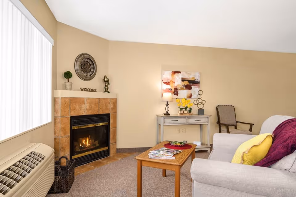 Cozy living room with a lit fireplace, sofa with colorful pillows, a coffee table and a side table with a lamp and decor.
