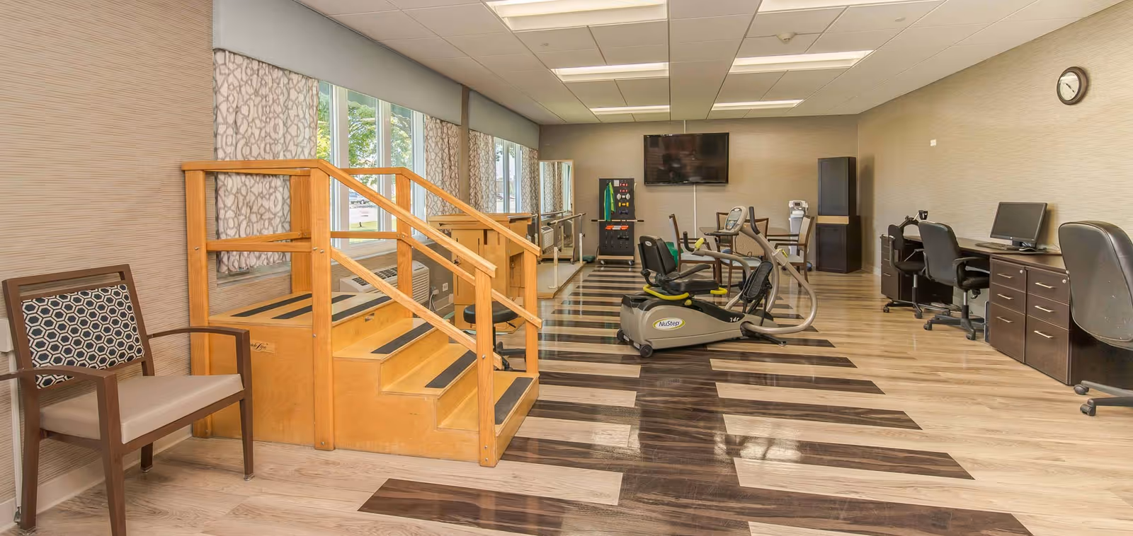 A rehabilitation or physical therapy room with wooden stairs for exercise, a recumbent exercise bike, parallel bars for walking practice, a vending machine, a wall-mounted TV, and desks with computers along the right wall. There is a chair with patterned upholstery on the left side near the stairs, and large windows with patterned curtains letting in natural light.