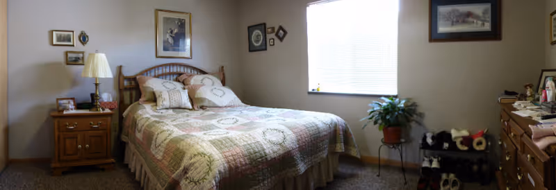 A cozy bedroom with a double bed covered in a patchwork quilt and several pillows. To the left of the bed is a wooden nightstand with a lamp, framed photos, and a small decorative item. The walls are adorned with various framed pictures. A window with blinds is on the right wall, letting in natural light. Below the window is a potted plant on a stand and a shoe rack with several pairs of shoes. On the right side of the room is a wooden dresser with various personal items on top.