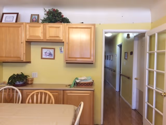 Interior view of a room with light wooden cabinets mounted on a yellow wall above a countertop. A table with a beige tablecloth and wooden chairs is in the foreground. To the right, there is a doorway leading to a hallway with handrails and framed pictures on the walls. The floor is wooden, and there is a glass-paneled door partially visible on the right side.