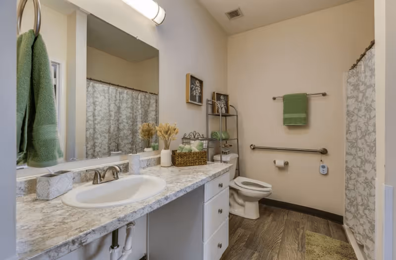 A clean bathroom with a marble countertop sink, a large mirror, and a green towel hanging on a ring. The bathroom has a toilet with a metal rack above it holding green towels and decorative items. There is a shower with a patterned curtain and a green bath mat on the wooden floor. The walls are light-colored and there are two framed floral pictures above the toilet.