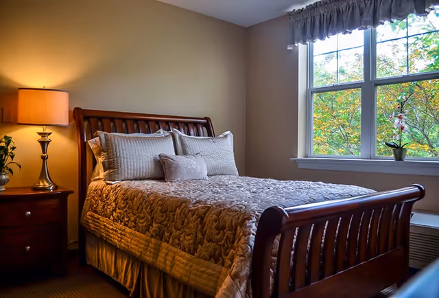 A cozy bedroom with a wooden bed frame, neatly made with a patterned quilt and multiple pillows. A wooden nightstand with a lamp and a small plant is beside the bed. A window with a valance shows green trees outside.