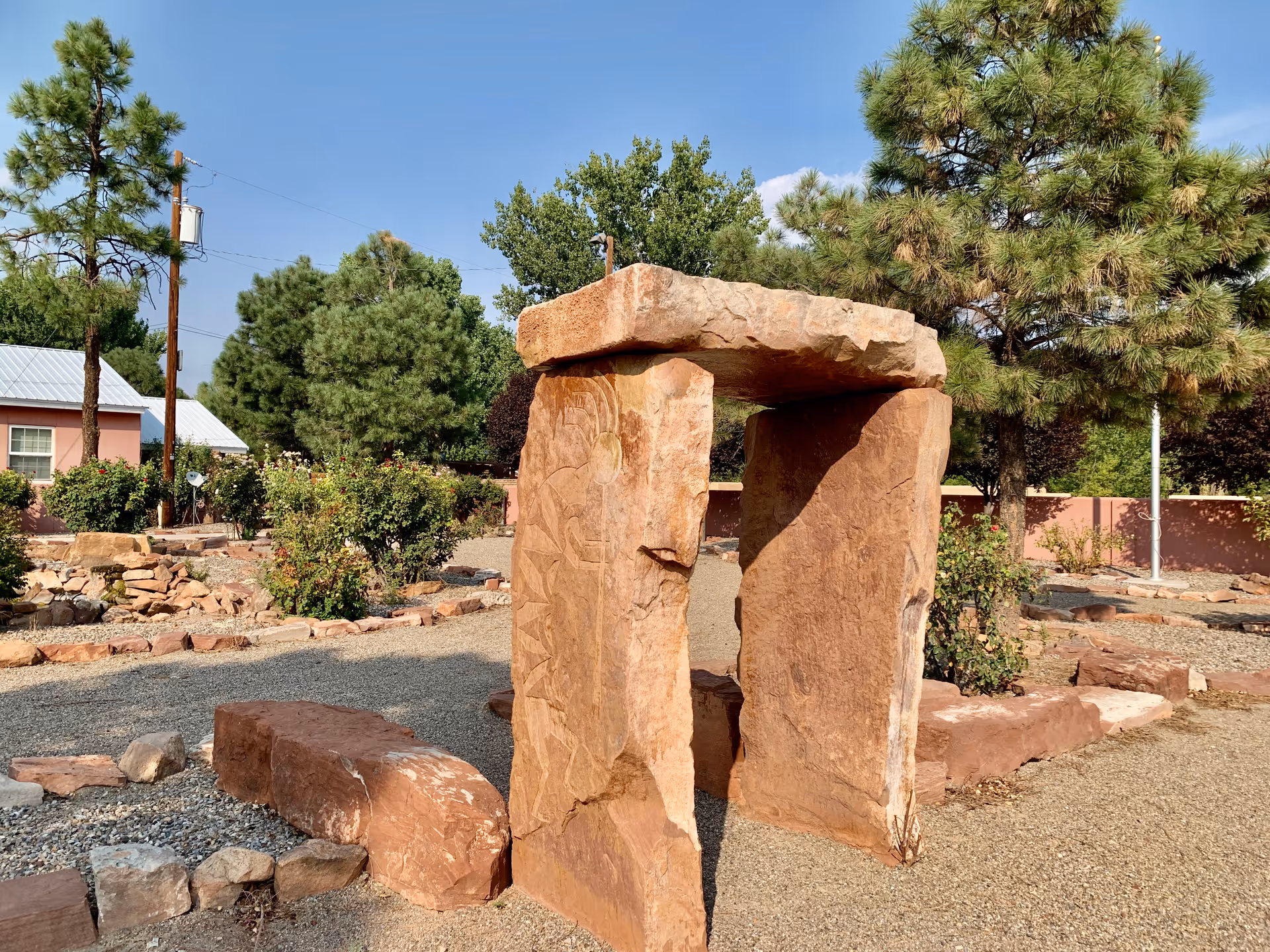 Outdoor garden area with a stone sculpture resembling a small stone arch or dolmen surrounded by gravel paths, bushes, and trees under a clear blue sky.