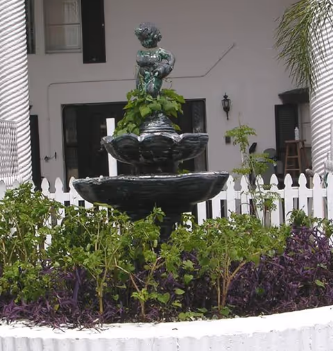 A black tiered water fountain with a small statue on top, surrounded by green and purple plants in a white circular planter. The background shows a white picket fence, white columns, and part of a building entrance with a door and windows.