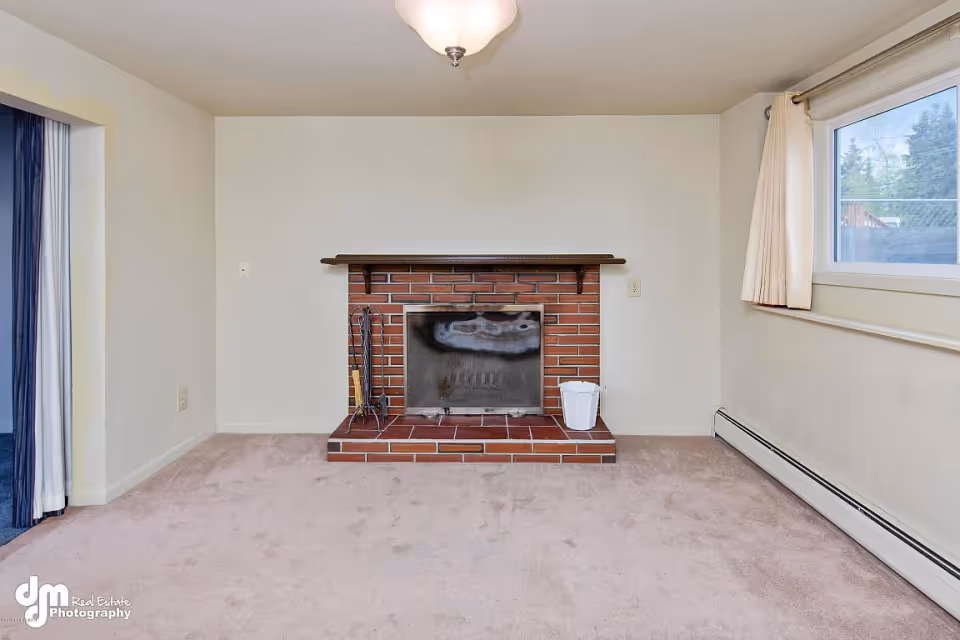 A small room with beige carpet and cream-colored walls featuring a brick fireplace with a wooden mantel. To the right of the fireplace is a white bucket, and to the left are fireplace tools. There is a window with cream curtains on the right wall, and an open doorway with dark blue curtains on the left. A ceiling light fixture is visible above.