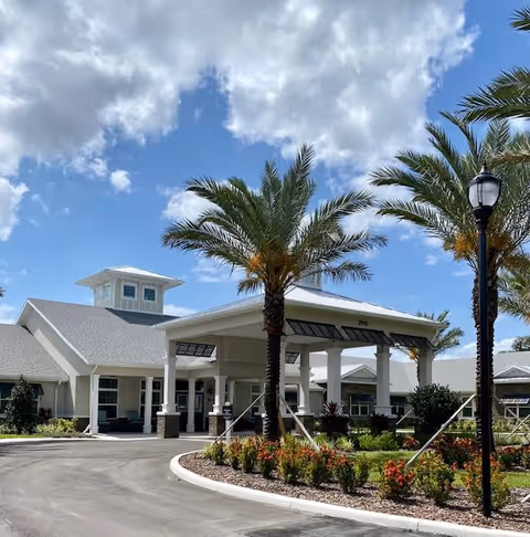 Covered porte-cochère entrance of a senior living building framed by palm trees, landscaped beds, and a partly cloudy sky.