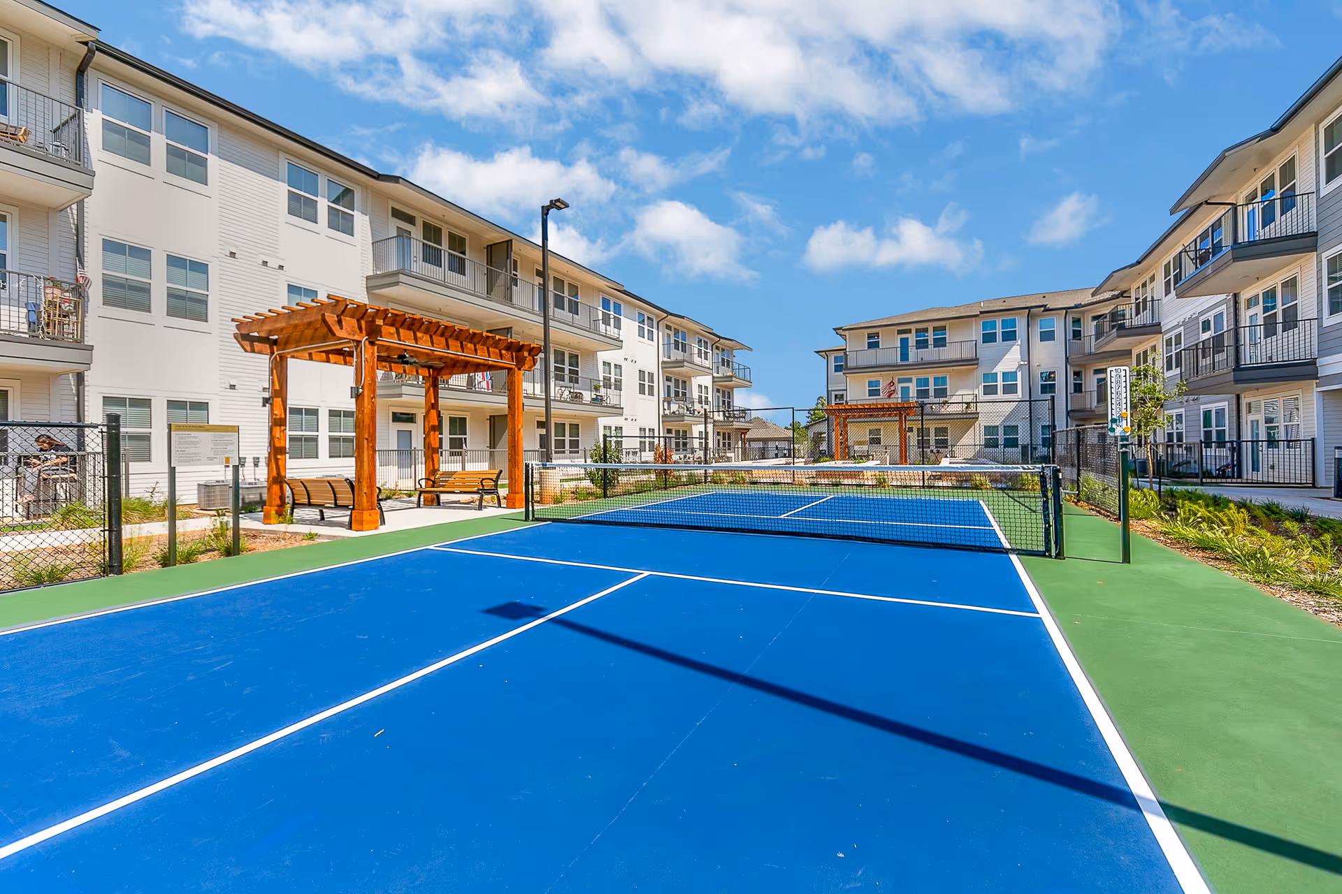 Blue outdoor pickleball/tennis court in a courtyard surrounded by three-story apartment buildings with wooden pergolas and benches.