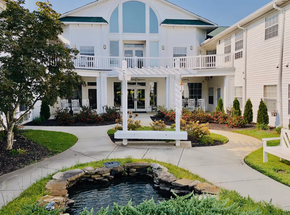 Front courtyard of a white two-story senior living building with a small pond, pergola bench, walkways, landscaping and a covered entrance with rocking chairs.