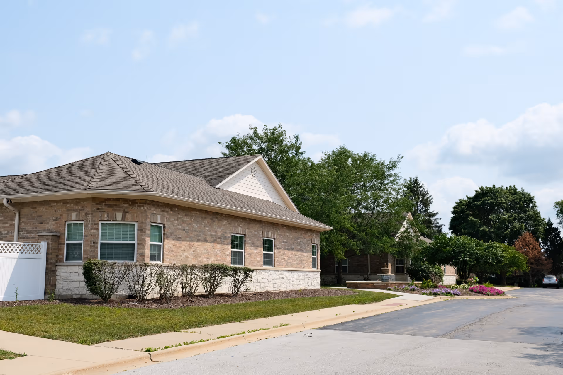 Exterior view of a single-story brick building with multiple windows, surrounded by trimmed bushes and trees under a partly cloudy sky. A paved road runs alongside the building with a sidewalk and landscaped flower beds.