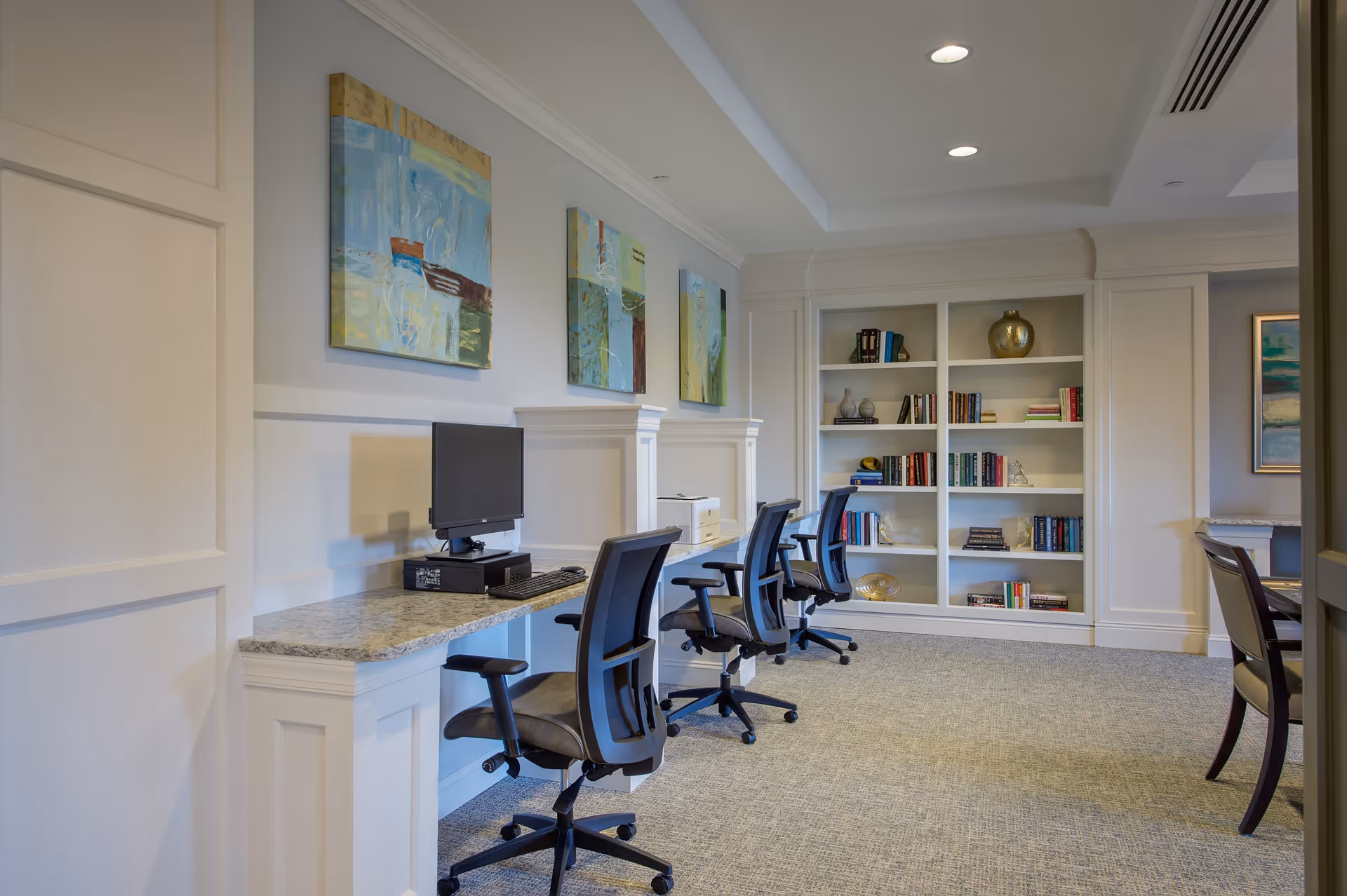 A quiet, well-lit room with a row of three black office chairs in front of a long granite countertop with desktop computers. The walls are decorated with abstract paintings, and there is a built-in white bookshelf filled with books and decorative items. The room has a carpeted floor and recessed ceiling lights.