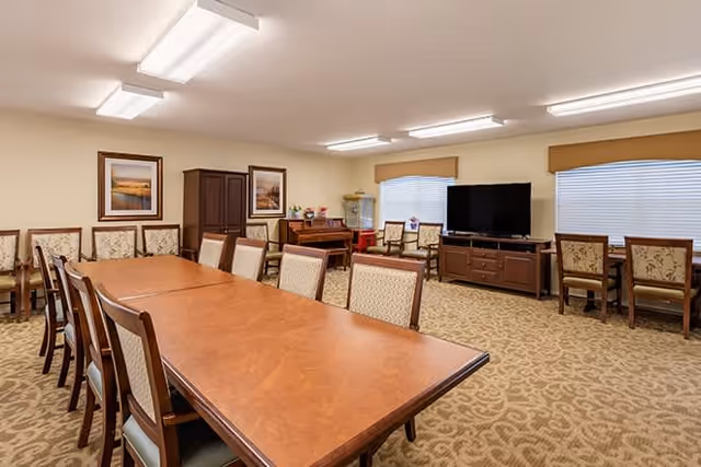 A spacious, well-lit common room with a long wooden table surrounded by chairs. The room features beige walls, carpeted floor with a patterned design, framed artwork on the walls, a piano, a large flat-screen TV on a wooden stand, and several additional chairs along the walls and near windows with blinds.