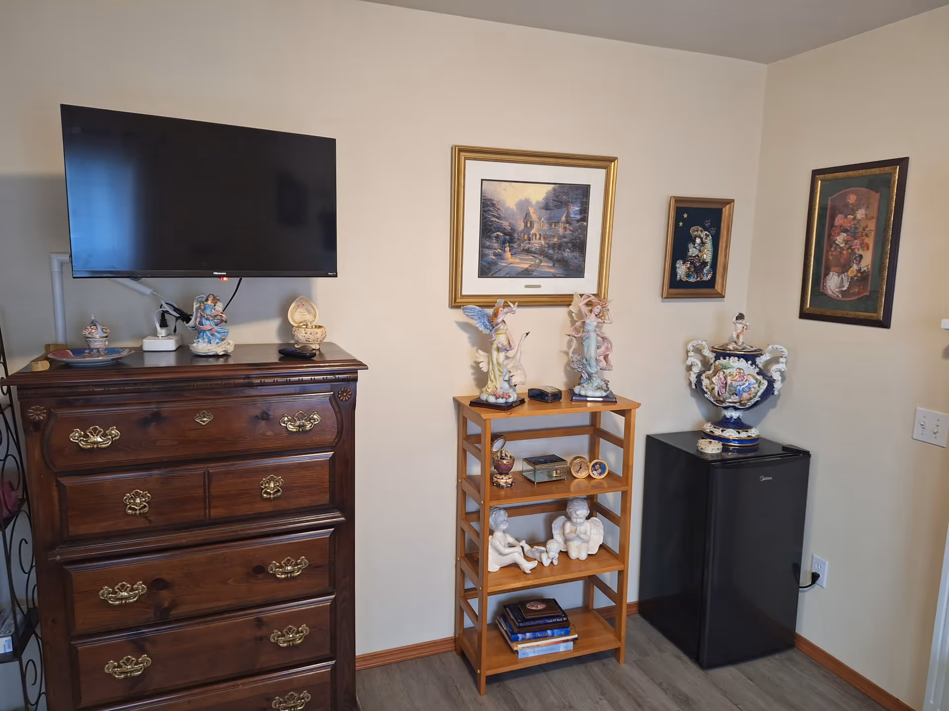 A corner of a furnished room with a wall-mounted TV above a wooden dresser, a small shelving unit displaying figurines and framed artwork, and a mini fridge.