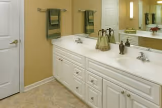 A clean and well-lit bathroom featuring a long white countertop with two sinks, silver faucets, and multiple drawers and cabinets underneath. Two green towels hang on the wall above the counter, and a large mirror spans the length of the countertop. The walls are painted a warm beige color, and the floor has light-colored tiles.