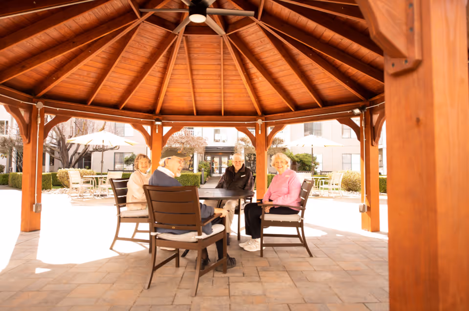 Four elderly people sitting around a table under a wooden gazebo in an outdoor courtyard area with patio furniture and umbrellas in the background.