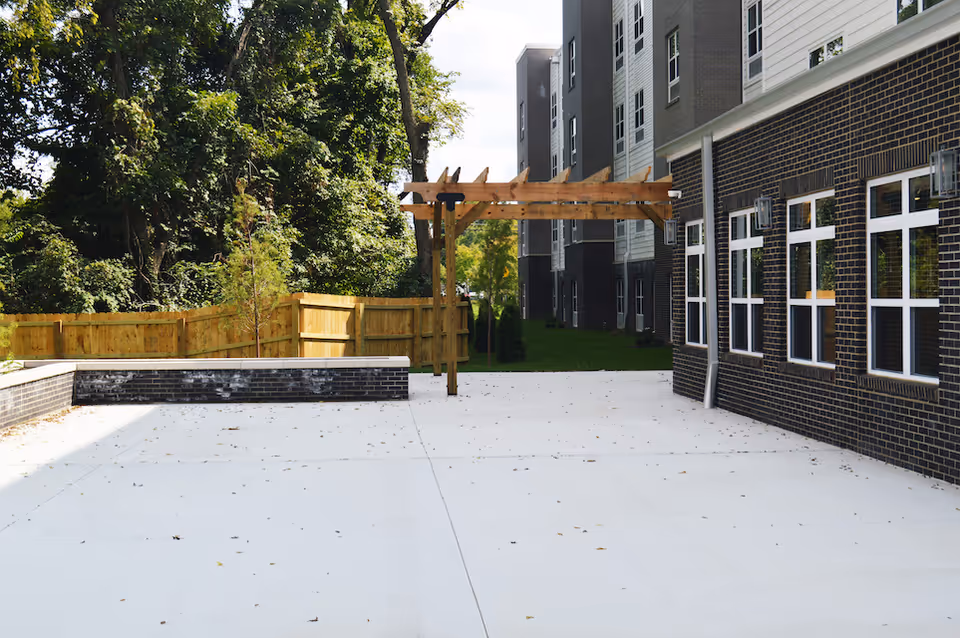 Outdoor patio area at Vivera Senior Living of Jeffersonville with a concrete floor, wooden pergola, brick building with multiple windows on the right, and a wooden fence with trees in the background.