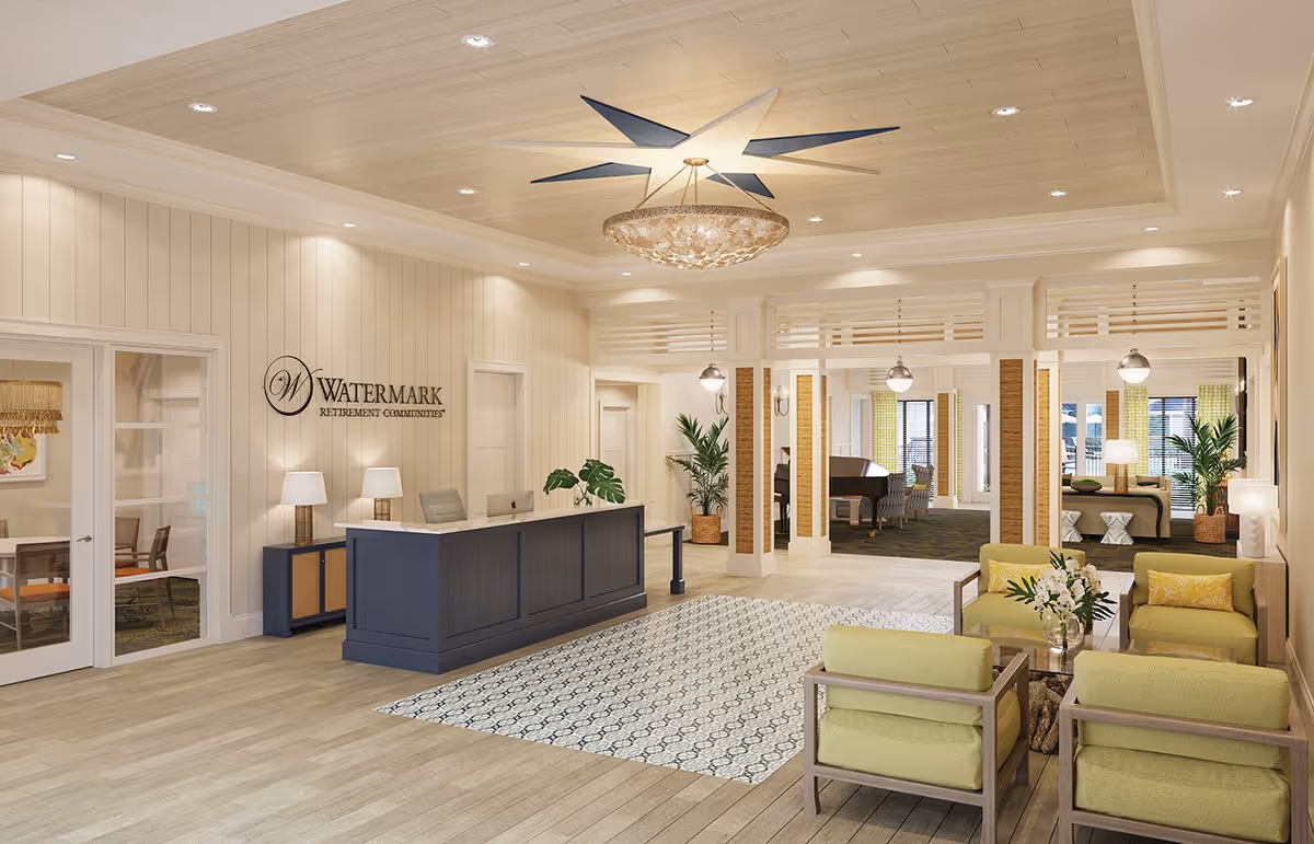 A bright and spacious reception area of a retirement community featuring a navy blue reception desk with a computer, two table lamps, and a plant. Behind the desk is a wall with the Watermark Retirement Communities logo. The room has light wood flooring, a patterned rug, and a ceiling with recessed lighting and a decorative chandelier. To the right, there is a seating area with four green cushioned chairs around a glass coffee table with a flower arrangement. In the background, there is a lounge area with a grand piano, additional seating, and large windows with green curtains.