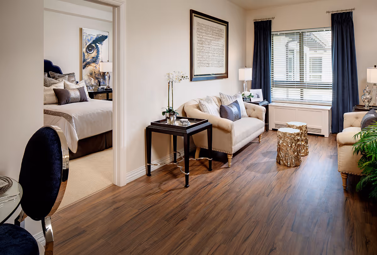 A cozy living room with two beige sofas adorned with decorative pillows, a small black side table with a white orchid, and two gold textured stools in the center. The room has wooden flooring, a large window with dark blue curtains, and a framed artwork on the wall. Through an open doorway, a bedroom with a bed, pillows, a nightstand, and a large owl painting is visible.