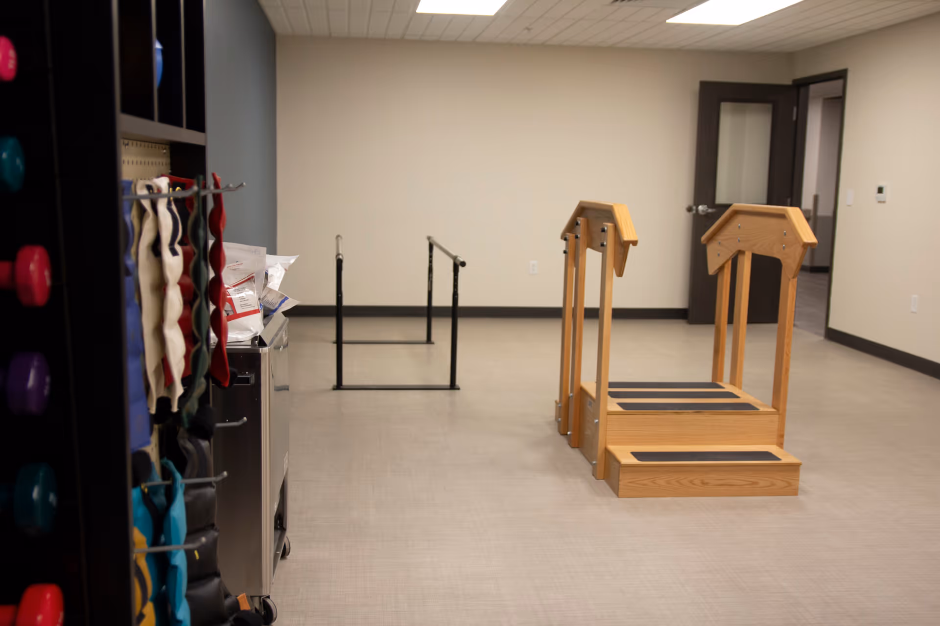 A rehabilitation or physical therapy room with wooden stairs equipped with handrails and parallel bars for walking exercises. On the left side, there is a rack holding various exercise equipment such as resistance bands and weights. The room has beige walls, a light-colored floor, and a dark door in the background.
