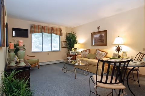 A cozy living room with a beige sofa adorned with patterned cushions, a glass coffee table with decorative items, a round dining table with chairs, a floor lamp, and a window with a plaid valance letting in natural light. There are plants and framed artwork on the walls, creating a warm and inviting atmosphere.