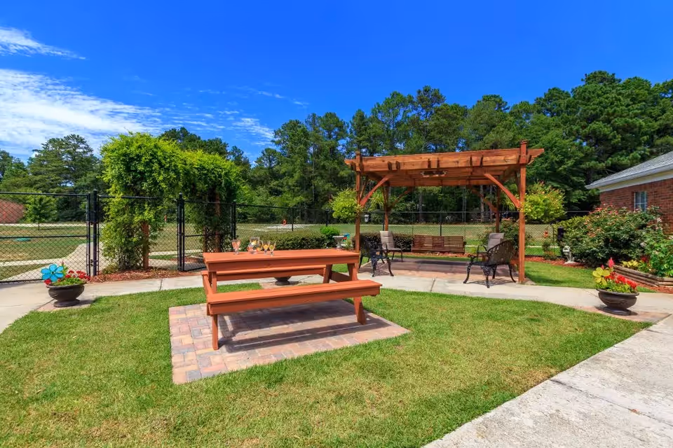 Outdoor garden area at Merryvale Assisted Living featuring a wooden picnic table with drinks on it, a pergola with seating including a swing and chairs, surrounded by green grass, potted plants, and trees under a bright blue sky.