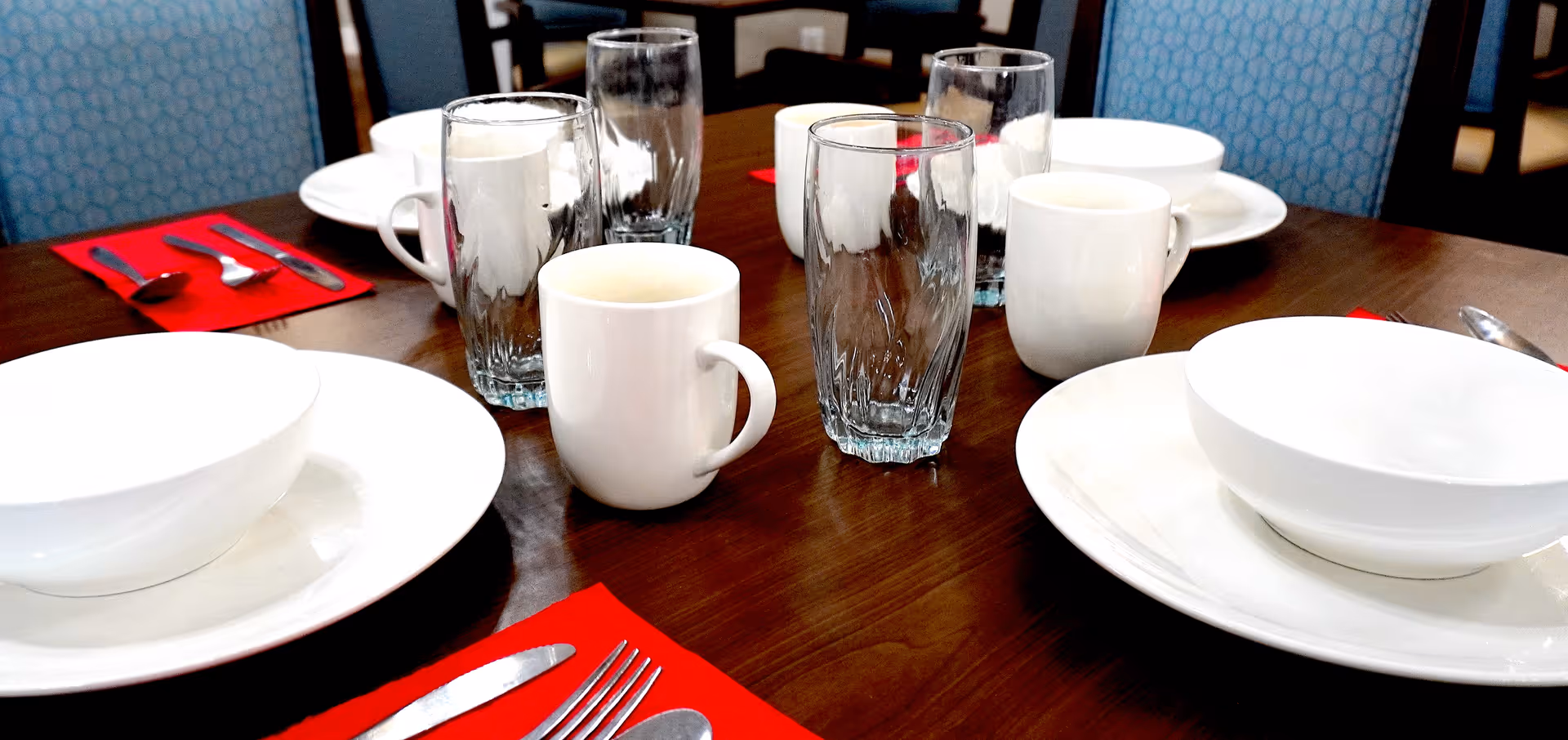 A dining table set with white bowls on white plates, white mugs, clear drinking glasses, and silverware placed on red napkins. Blue patterned chairs are visible around the table.