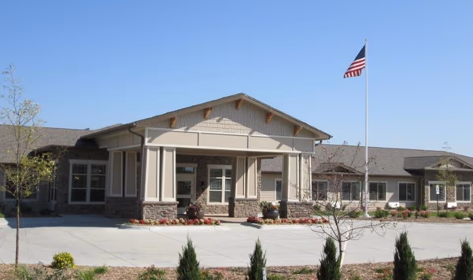 Exterior view of Cedar Creek of High Plains senior living facility showing a single-story building with a covered entrance, stone and beige siding, an American flag on a flagpole, and landscaped grounds under a clear blue sky.