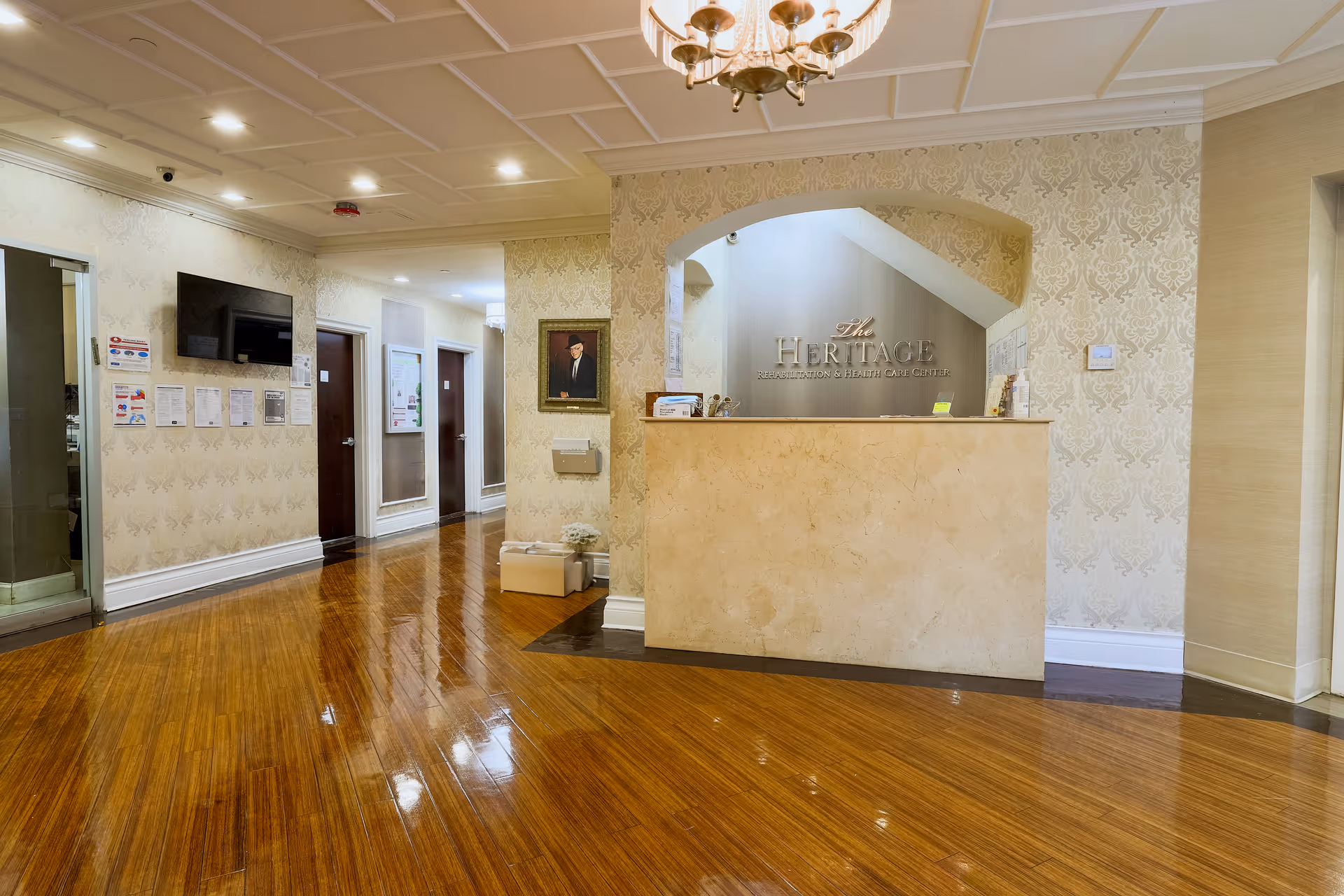 Reception area of Heritage Center for Rehabilitation with a beige marble front desk, patterned wallpaper, wooden floor, a chandelier on the ceiling, a portrait on the wall, and a hallway with doors and a wall-mounted TV.