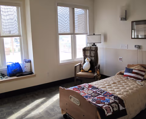A bright bedroom with a single bed covered in a beige quilt and a colorful patchwork blanket. There is a pillow with an American flag design on the bed. Next to the bed is a wooden armchair with a cushion and a stuffed penguin toy sitting on it. The room has two large windows with white blinds partially drawn, letting in natural light. A small table lamp is mounted on the wall above the chair. The floor has a patterned carpet, and there are some bags placed on the window sill.