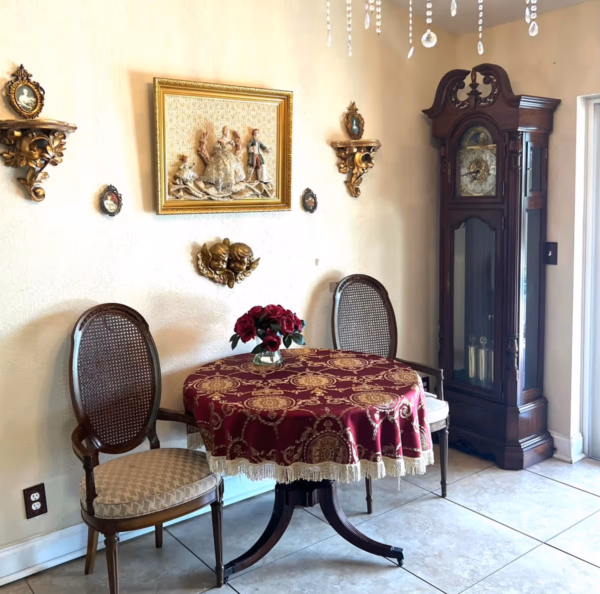 A small round table covered with a red and gold patterned tablecloth with fringe, holding a vase of red roses. Two wooden chairs with cushioned seats and cane backs are placed on either side of the table. On the wall behind the table are decorative shelves with small framed portraits, a framed relief artwork of three figures, and a pair of cherub wall sculptures. To the right stands a tall, dark wooden grandfather clock. The floor is tiled, and part of a glass door is visible on the right.
