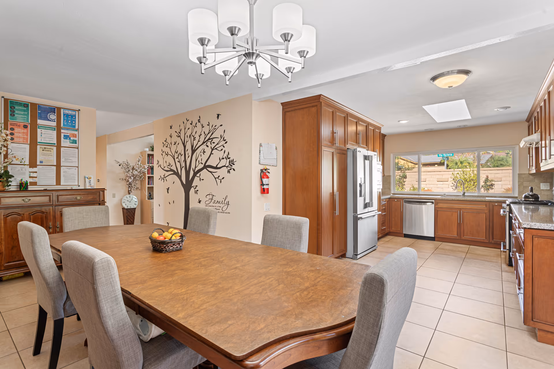 A spacious kitchen and dining area with a large wooden dining table surrounded by six upholstered chairs. The kitchen features wooden cabinets, stainless steel appliances including a refrigerator, dishwasher, and stove, and a large window above the sink letting in natural light. A decorative tree wall decal with the word 'Family' is on the wall near the dining area, and a chandelier hangs above the table.