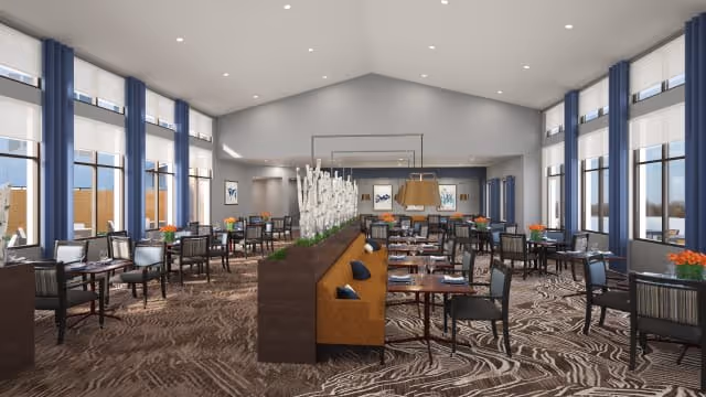 Spacious dining room with multiple tables and chairs arranged neatly. The room features large windows with white blinds, high ceilings with recessed lighting, and a central divider with decorative plants. The carpet has a patterned design, and there are framed artworks on the walls.