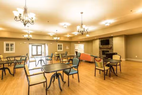 A spacious senior living common area with multiple tables, chairs, chandeliers, and a fireplace.