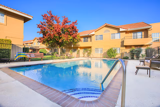 A sunny courtyard with a rectangular swimming pool, lounge chairs, and a two-story tan building with balconies and a red-leaf tree.