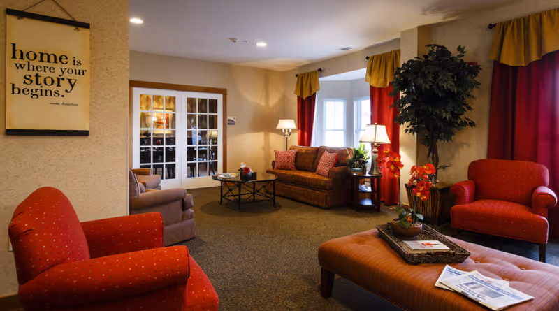 A cozy living room area with red and patterned armchairs, a patterned sofa, a large ottoman with a tray and newspapers, a coffee table, lamps, a large potted plant, and windows with red and yellow curtains. A framed wall art with the quote 'home is where your story begins.' is visible on the left wall.