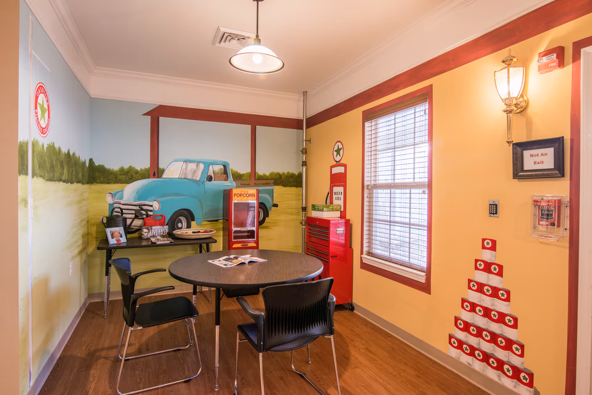 A small room with a round table and four chairs. The walls are painted with a mural of a blue vintage pickup truck in a countryside setting. There is a popcorn machine, a red toolbox, and a window with blinds. The wall also features a painted vintage gas pump and a pyramid made of red and white boxes. A light fixture hangs from the ceiling, and a wall sconce is mounted near a sign that reads 'Not An Exit.'
