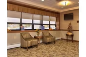A waiting area with two striped armchairs and a small round wooden table between them, placed in front of a large window with blinds. The floor is carpeted with a patterned design, and the walls are decorated with a mounted flat-screen TV and a small table holding a decorative item. The ceiling has recessed lighting and a central light fixture.